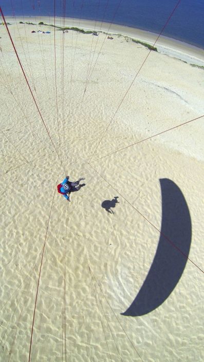 Flugaufnahme mit Kameramontage im Untersegel: Man sieht die Gleitschirmleinen und den Piloten bodennah fliegend sowie seinen Schatten.