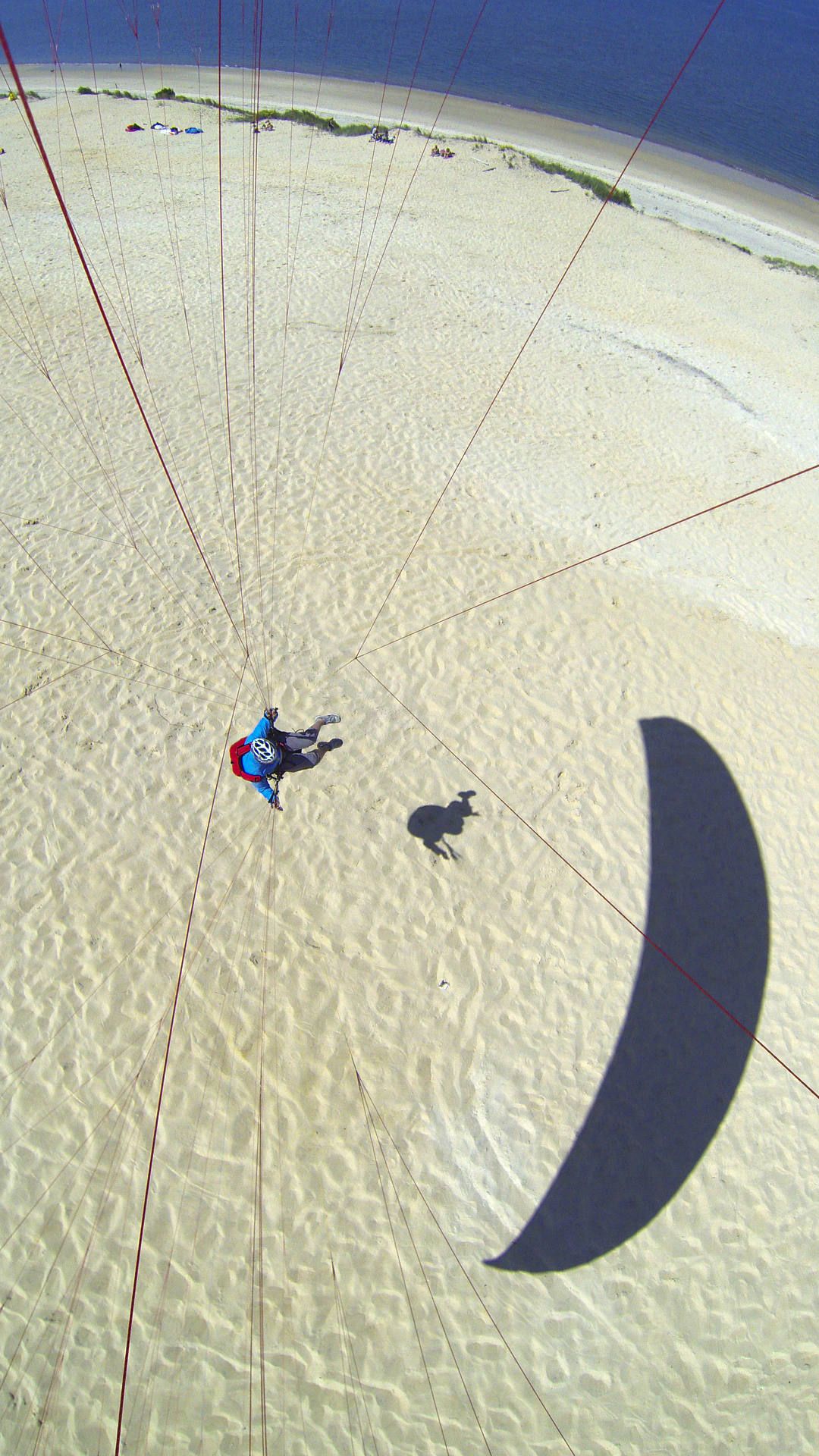 Flugaufnahme mit Kameramontage im Untersegel: Man sieht die Gleitschirmleinen und den Piloten bodennah fliegend sowie seinen Schatten.