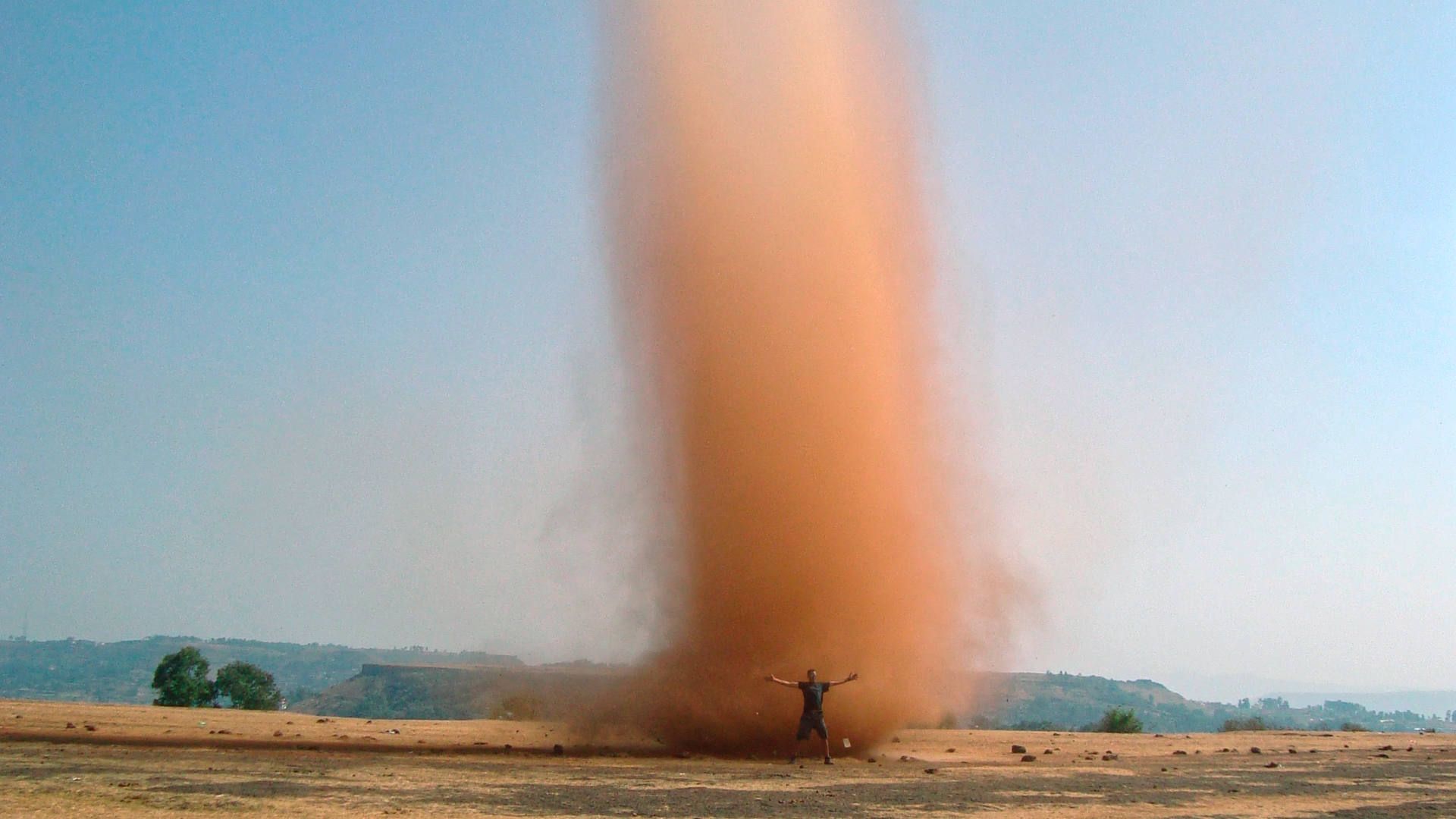 Ein Gleitschirmpilot wagt sich nahe an einen Dust Devil heran und zeigt mit ausgestreckten Armen das Grössenverhältnis.